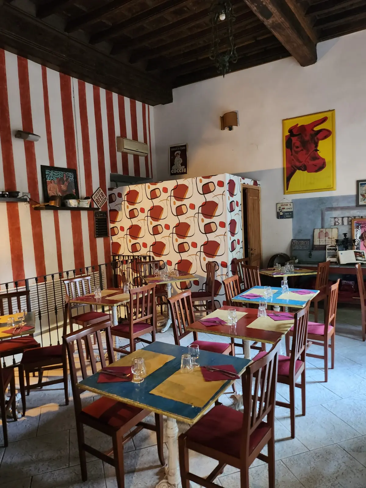 La sala: parete a righe rosse e bianche, tavoli gialli e blu, travi di legno